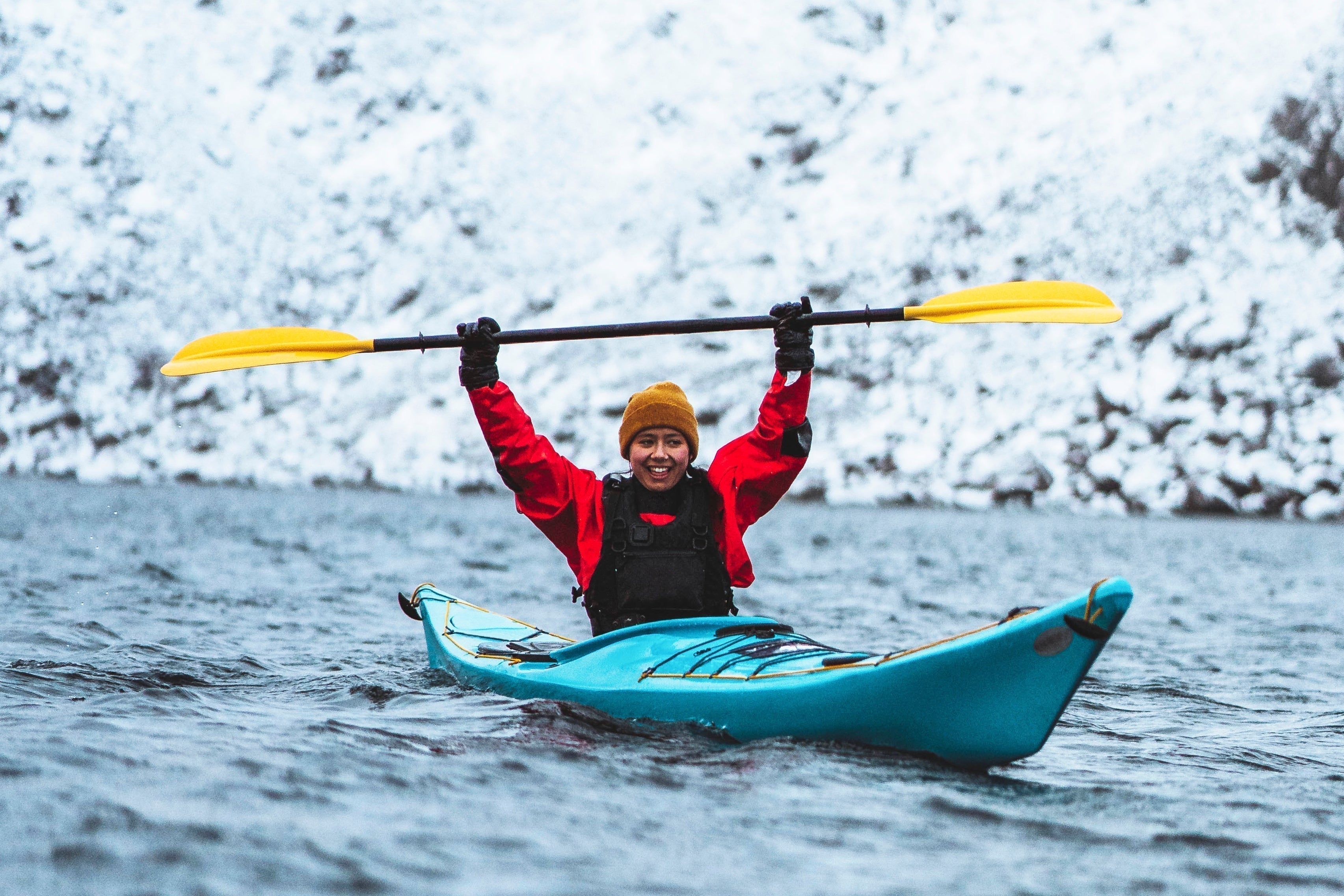 Woman kayaking in cold water wearing a red drysuit and yellow beanie, snowy fjord background